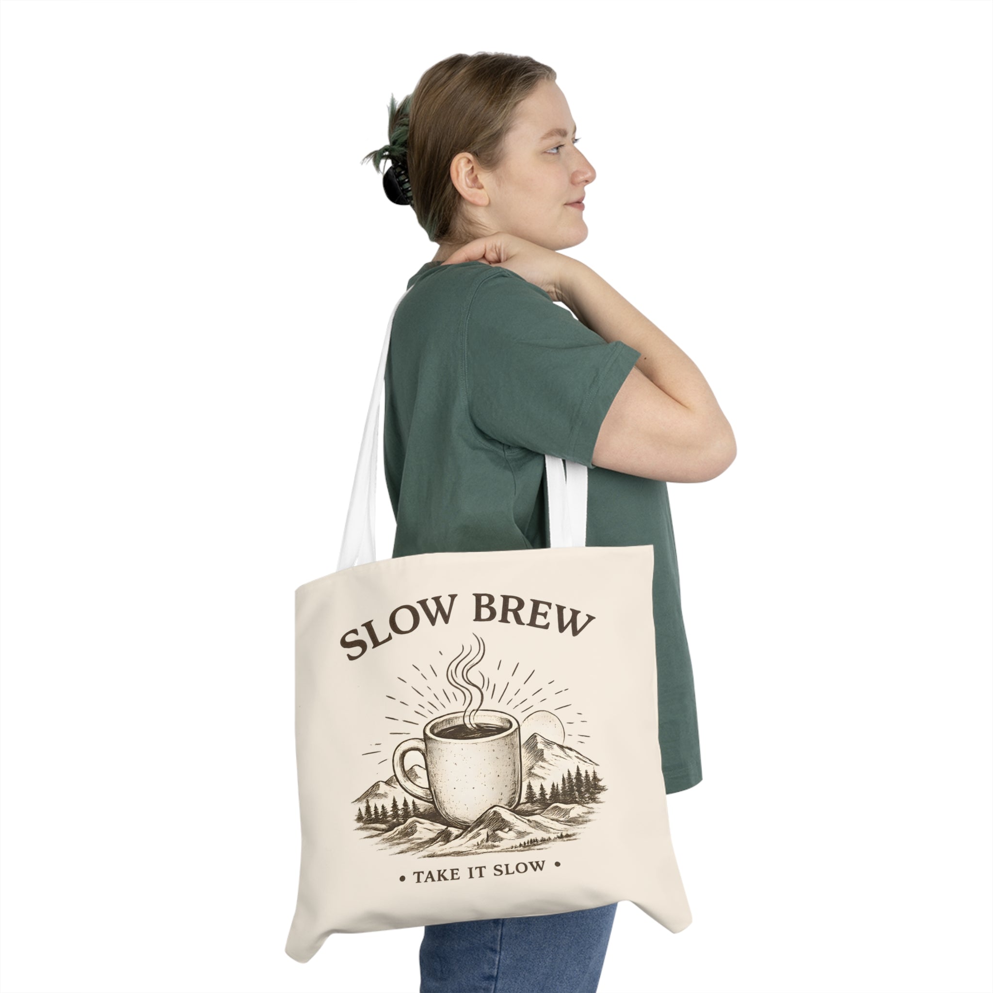 A woman wearing a dark green t-shirt and blue jeans is shown from the back and side, carrying a cream canvas tote bag over her shoulder. The bag features a brown graphic of a steaming coffee mug with mountains and trees, and the text 'SLOW BREW' and 'TAKE IT SLOW'.