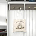 A beige canvas tote bag with white shoulder straps hangs from a wooden coat rack hook in a bright, modern entryway. The bag features a dark brown graphic of a steaming coffee mug with the text 'SLOW BREW' and 'TAKE IT SLOW'.